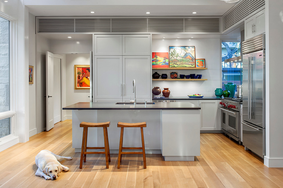 Photo of a mid-sized transitional l-shaped eat-in kitchen in Austin with an undermount sink, recessed-panel cabinets, grey cabinets, solid surface benchtops, white splashback, subway tile splashback, stainless steel appliances, light hardwood floors and with island.