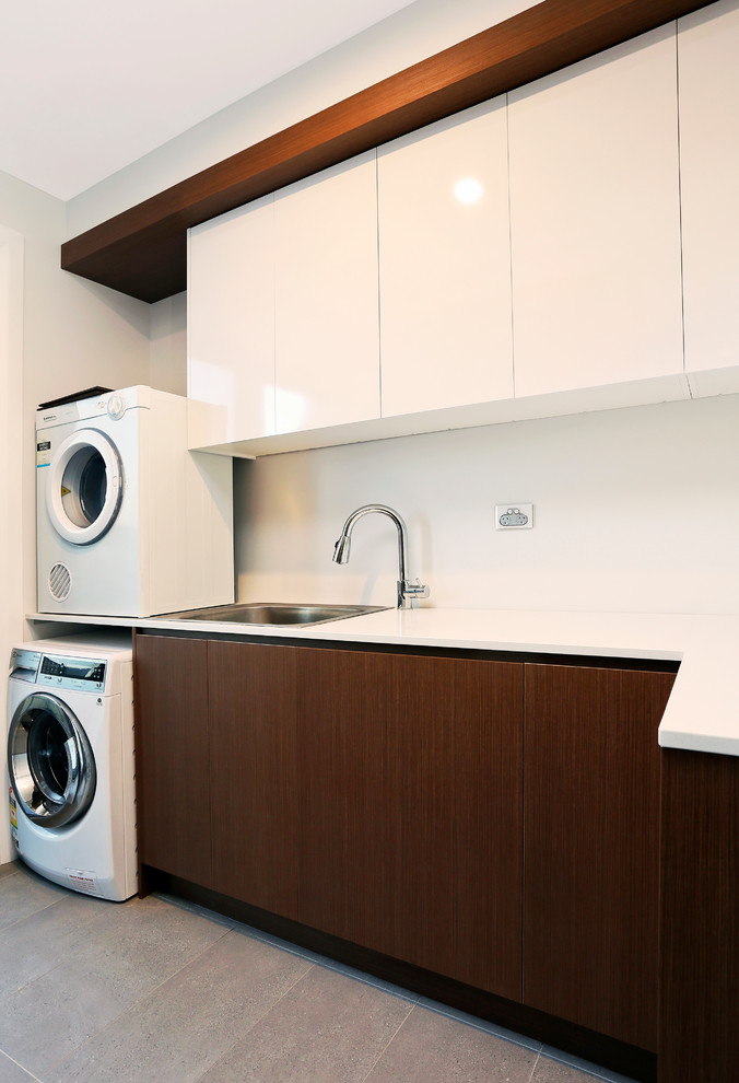 Inspiration for a large contemporary l-shaped dedicated laundry room in Sydney with flat-panel cabinets, white cabinets, quartz benchtops, metallic splashback, mirror splashback, porcelain floors, grey floor, white benchtop, a drop-in sink, white walls and a stacked washer and dryer.