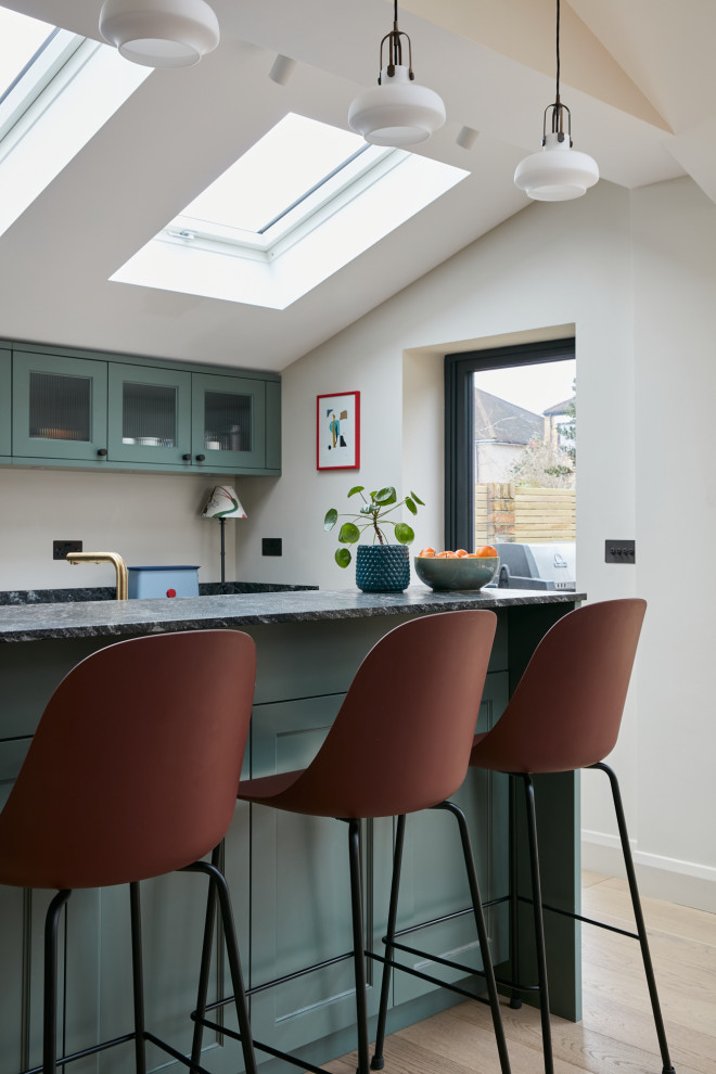 This is an example of a mid-sized traditional l-shaped open plan kitchen in London with a farmhouse sink, recessed-panel cabinets, green cabinets, granite benchtops, black splashback, granite splashback, light hardwood floors, with island, black benchtop and vaulted.