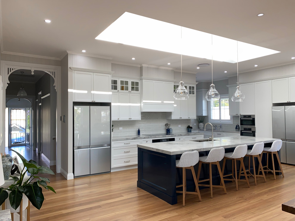 Photo of an expansive contemporary l-shaped open plan kitchen in Brisbane with a double-bowl sink, recessed-panel cabinets, white cabinets, marble benchtops, grey splashback, subway tile splashback, stainless steel appliances, medium hardwood floors, with island, brown floor and white benchtop.