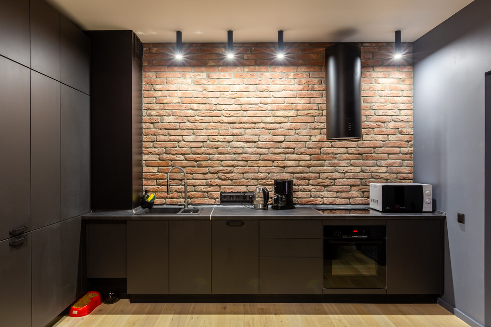 This is an example of an industrial single-wall kitchen in Moscow with a drop-in sink, flat-panel cabinets, black cabinets, brick splashback, black appliances, light hardwood floors, beige floor and black benchtop.