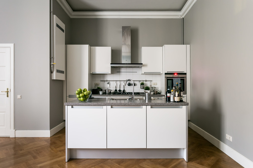 Photo of a contemporary galley kitchen in Saint Petersburg with an undermount sink, flat-panel cabinets, white cabinets, white splashback, stainless steel appliances, medium hardwood floors, with island, brown floor and grey benchtop.