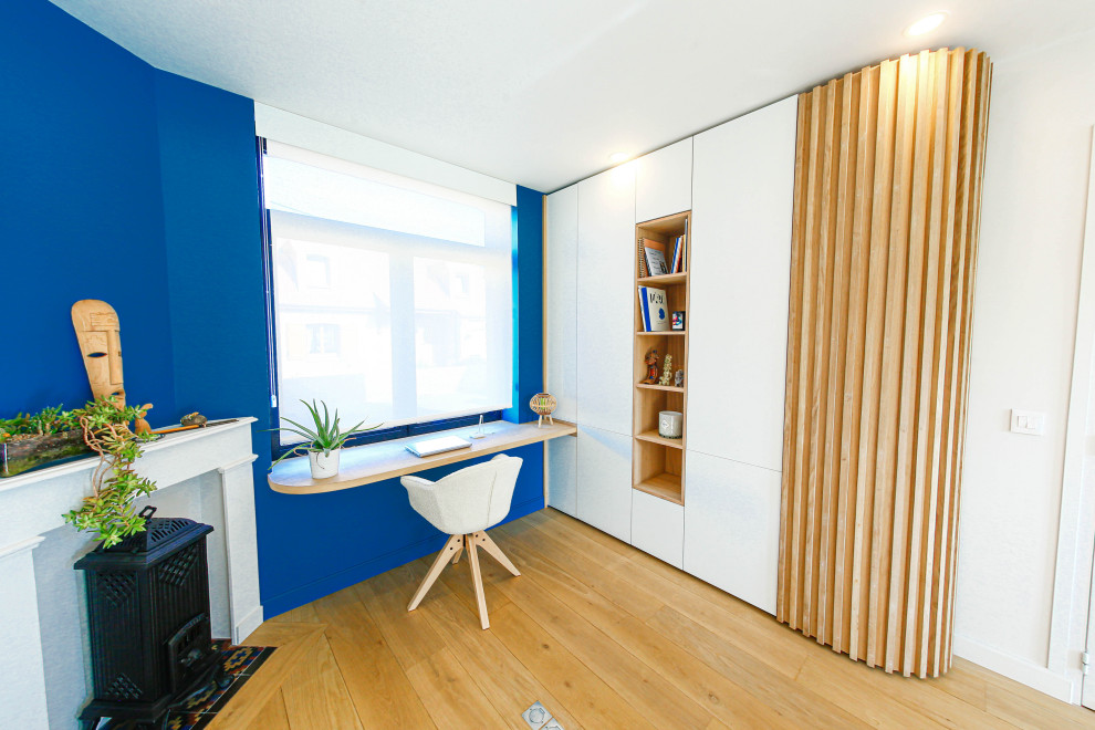 Medium sized study in Lille with blue walls, light hardwood flooring, a standard fireplace, a stone fireplace surround, a built-in desk and beige floors.