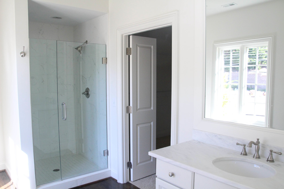 Photo of a large transitional master bathroom in Birmingham with flat-panel cabinets, white cabinets, a drop-in tub, a corner shower, a two-piece toilet, white tile, ceramic tile, white walls, dark hardwood floors, a drop-in sink, marble benchtops, brown floor, a hinged shower door and white benchtops.