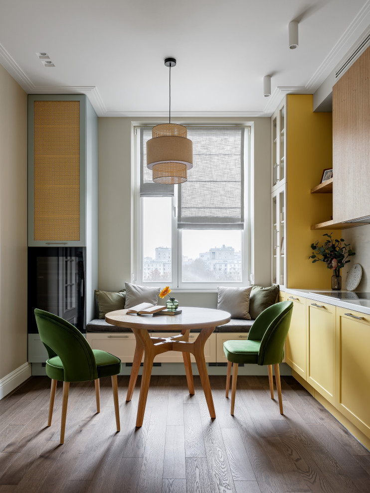 Inspiration for a mid-sized transitional single-wall kitchen in Saint Petersburg with an undermount sink, beaded inset cabinets, yellow cabinets, solid surface benchtops, white splashback, engineered quartz splashback, black appliances, medium hardwood floors, brown floor and white benchtop.