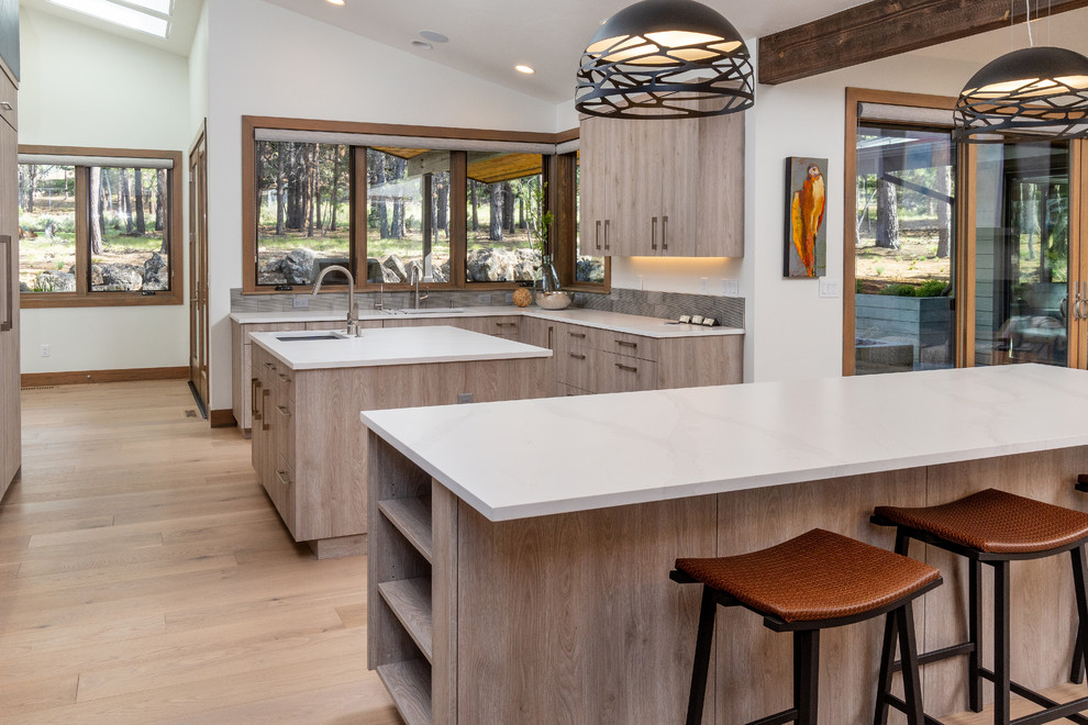 Example of a large mountain style u-shaped light wood floor open concept kitchen design with an undermount sink, flat-panel cabinets, light wood cabinets, quartz countertops, gray backsplash, ceramic backsplash, paneled appliances, two islands and white countertops