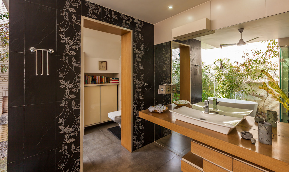 Photo of a contemporary bathroom in Delhi with a vessel sink, flat-panel cabinets, medium wood cabinets, a freestanding tub and black tile.