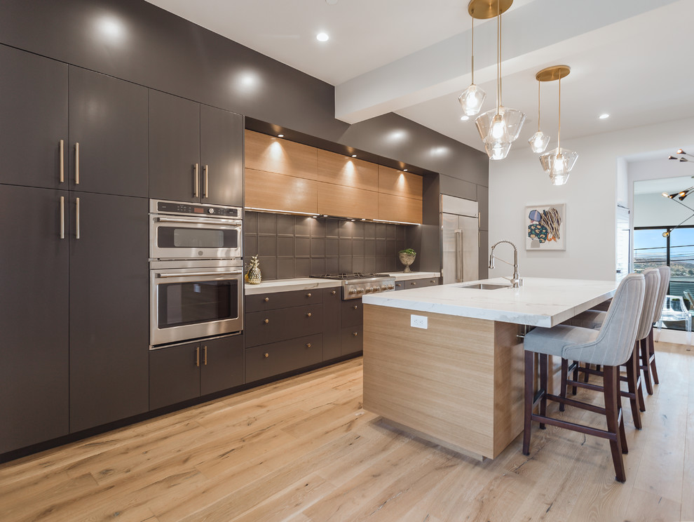 This is an example of a large midcentury single-wall open plan kitchen in Los Angeles with flat-panel cabinets, grey cabinets, grey splashback, glass tile splashback, stainless steel appliances, light hardwood floors, with island and white benchtop.