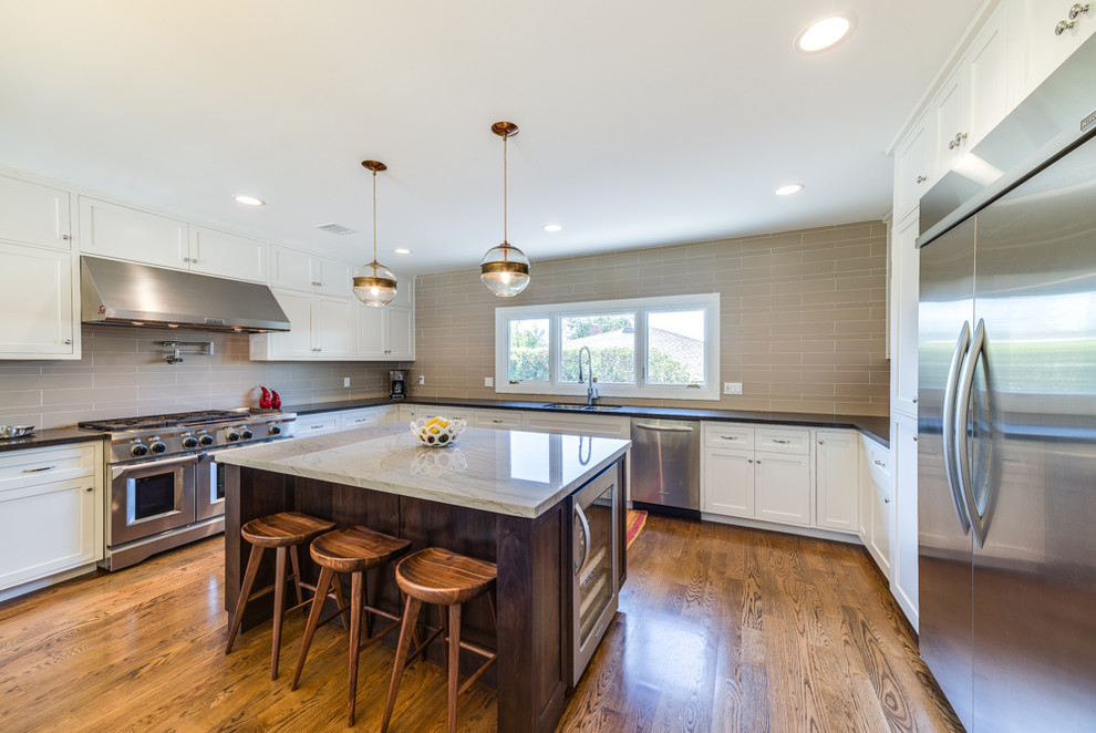 Photo of a large transitional u-shaped eat-in kitchen in Los Angeles with shaker cabinets, white cabinets, beige splashback, stainless steel appliances, medium hardwood floors, with island, a double-bowl sink, ceramic splashback, marble benchtops and brown floor.