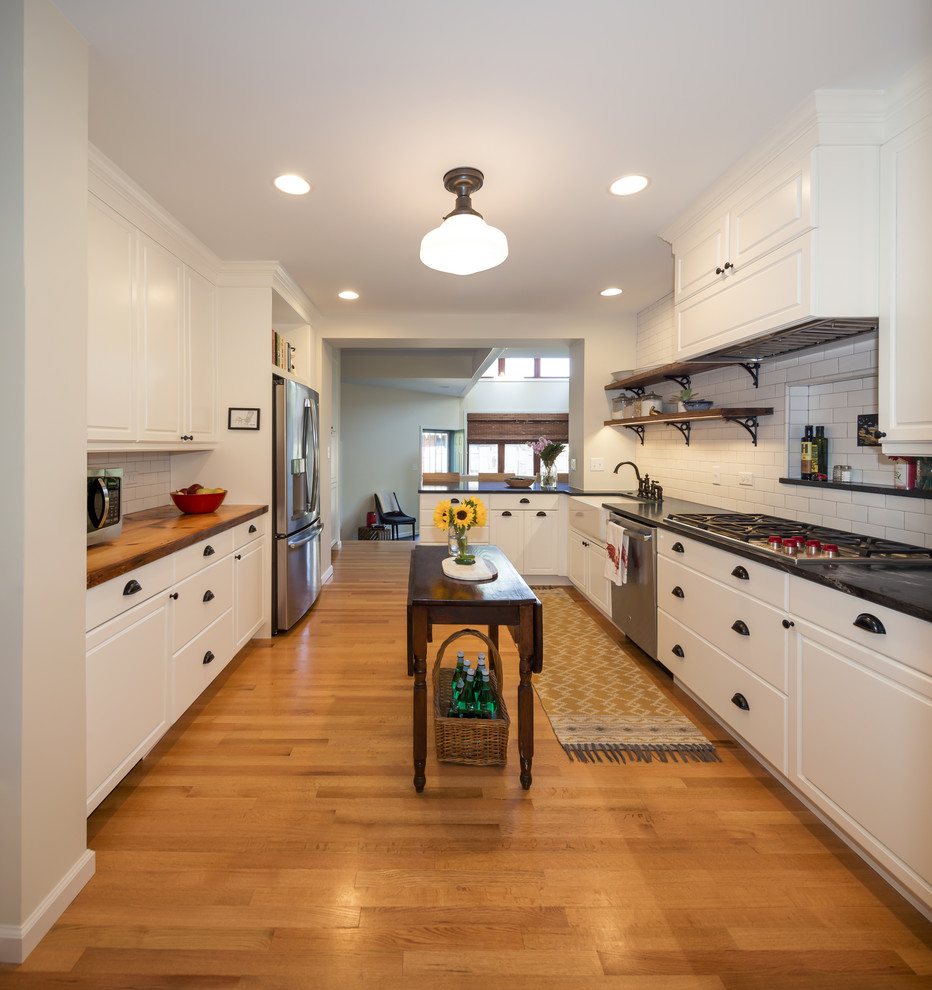 Large country l-shaped separate kitchen in Denver with white cabinets, white splashback, ceramic splashback, medium hardwood floors, a peninsula, a farmhouse sink, stainless steel appliances, shaker cabinets and black benchtop.