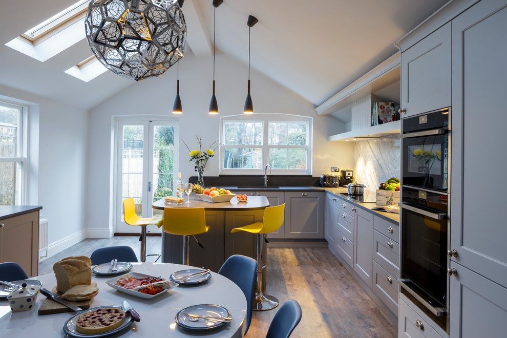 This is an example of a traditional open plan kitchen in Berkshire with a double-bowl sink, shaker cabinets, grey cabinets, quartzite benchtops, marble splashback, black appliances, with island, white splashback, vinyl floors and brown floor.
