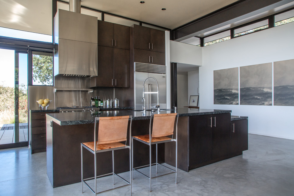 This is an example of a contemporary kitchen in Santa Barbara with an undermount sink, flat-panel cabinets, dark wood cabinets, metallic splashback, stainless steel appliances, concrete floors and with island.