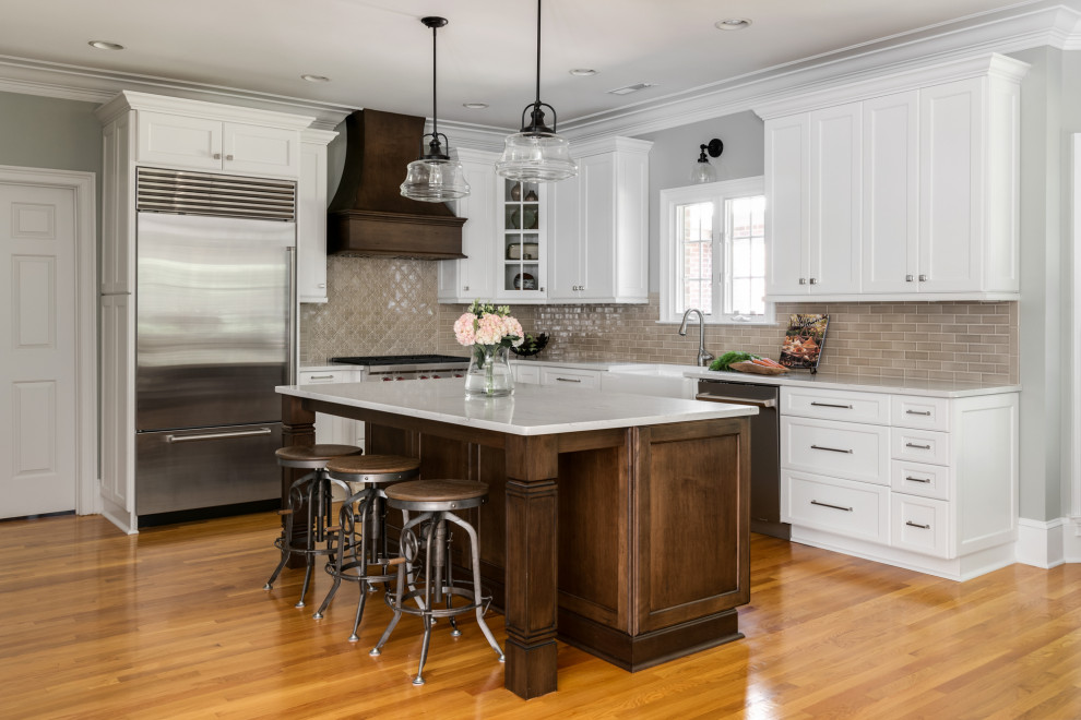 Photo of a mid-sized traditional l-shaped kitchen in Charlotte with a farmhouse sink, shaker cabinets, white cabinets, quartz benchtops, brown splashback, porcelain splashback, stainless steel appliances, light hardwood floors, with island, brown floor and grey benchtop.
