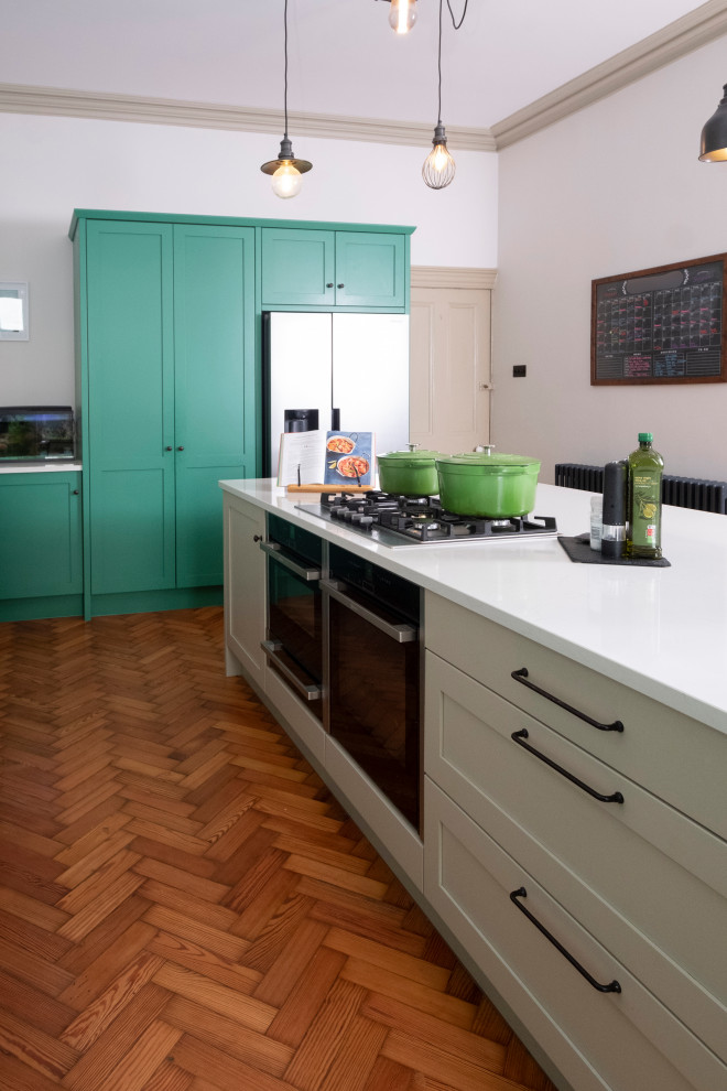 Photo of a large bohemian l-shaped kitchen/diner in Other with shaker cabinets, green cabinets, quartz worktops, white splashback, engineered quartz splashback, integrated appliances, light hardwood flooring, an island and white worktops.