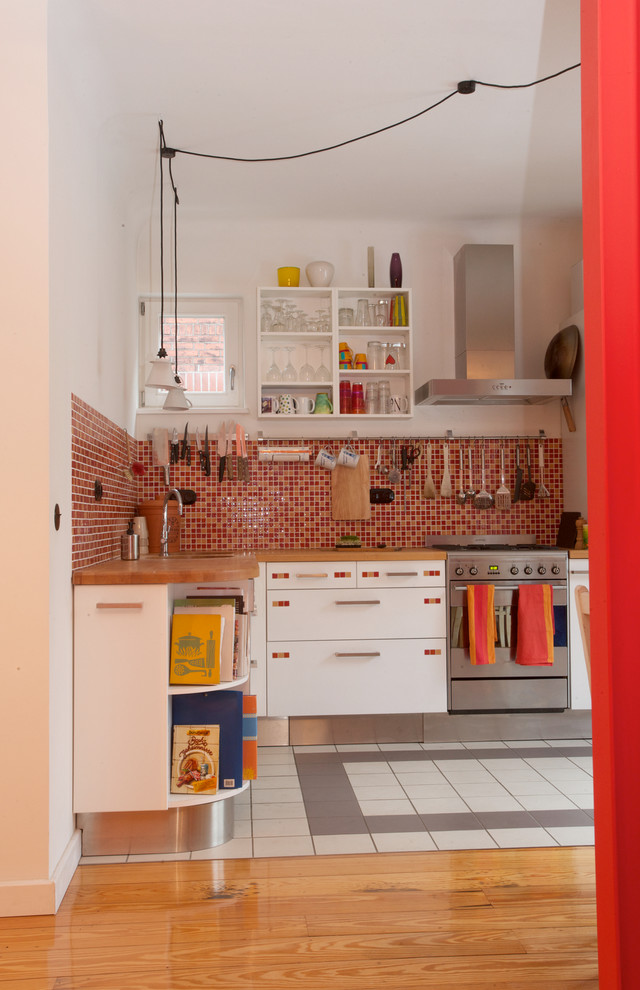 Photo of a mid-sized eclectic l-shaped open plan kitchen in Hamburg with flat-panel cabinets, white cabinets, red splashback, stainless steel appliances, no island, wood benchtops, a double-bowl sink, glass tile splashback, cement tiles and grey floor.