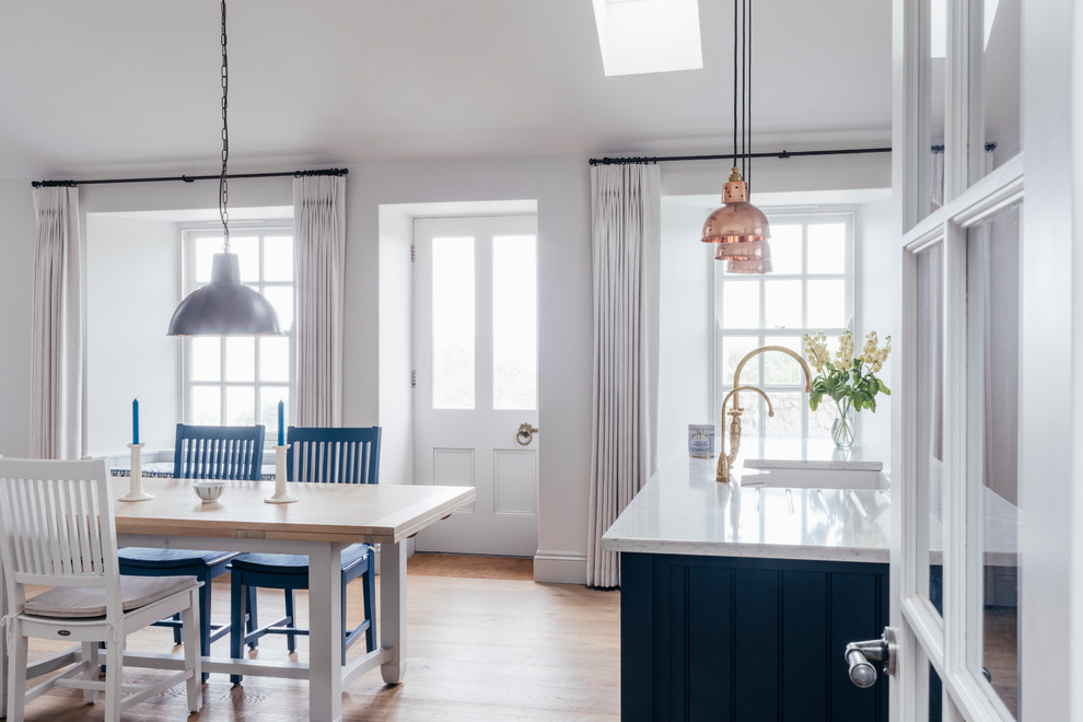 Mid-sized beach style single-wall open plan kitchen in Cornwall with a farmhouse sink, shaker cabinets, grey cabinets, solid surface benchtops, blue splashback, ceramic splashback, stainless steel appliances, medium hardwood floors, with island, brown floor and white benchtop.