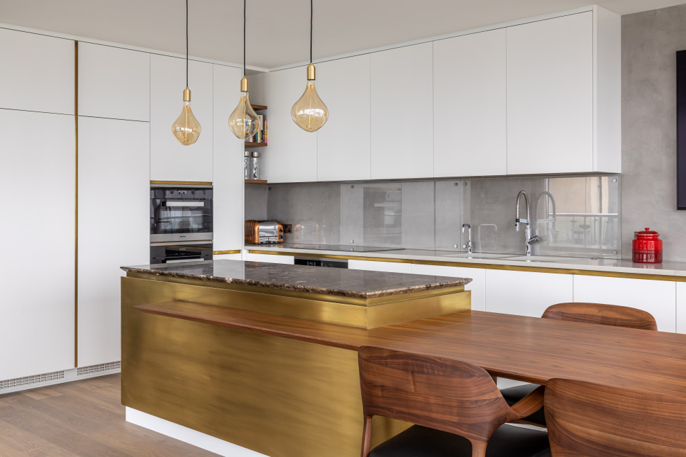 Large contemporary l-shaped kitchen in London with a double-bowl sink, flat-panel cabinets, white cabinets, grey splashback, glass sheet splashback, stainless steel appliances, medium hardwood floors, with island, brown floor and white benchtop.