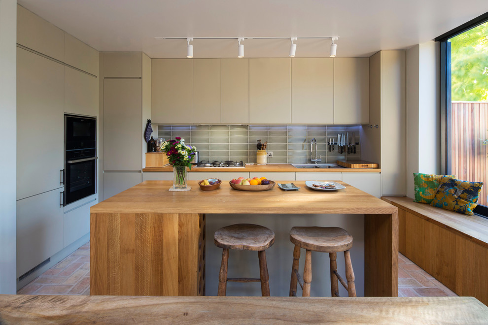 Inspiration for a transitional l-shaped eat-in kitchen in London with a double-bowl sink, flat-panel cabinets, beige cabinets, wood benchtops, grey splashback, glass tile splashback, black appliances, brick floors, with island, red floor and brown benchtop.