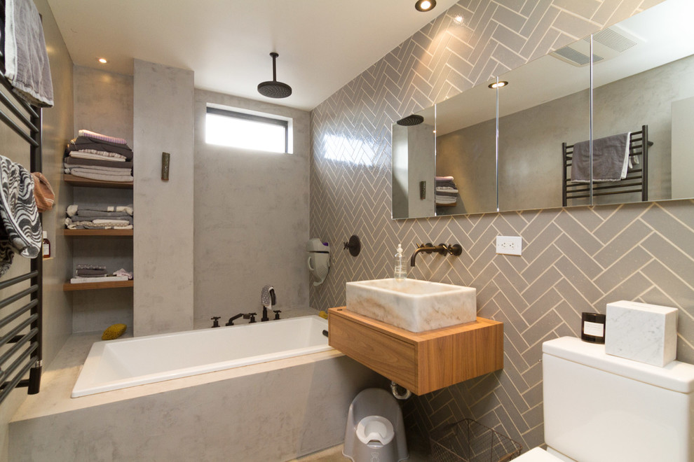 Contemporary bathroom in New York with a vessel sink, flat-panel cabinets, light wood cabinets, wood benchtops, a drop-in tub, a shower/bathtub combo, a two-piece toilet, gray tile, ceramic tile and grey walls.
