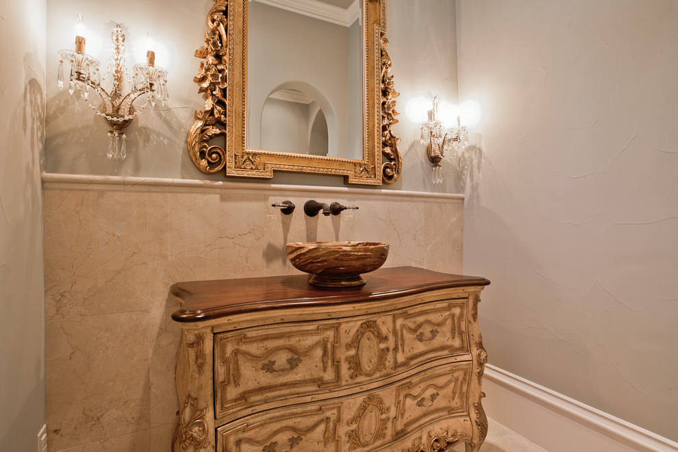 Inspiration for a mid-sized traditional powder room in Dallas with beaded inset cabinets, beige cabinets, a two-piece toilet, beige tile, stone tile, beige walls, porcelain floors, a vessel sink, wood benchtops, beige floor and brown benchtops.