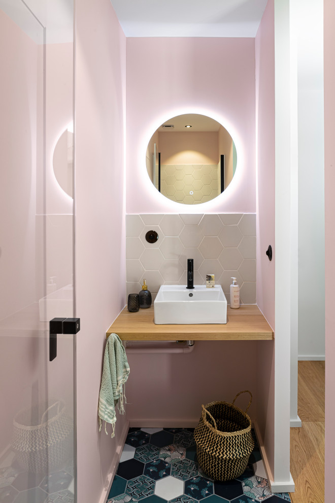 На фото: туалет в современном стиле с серой плиткой, розовыми стенами, настольной раковиной, столешницей из дерева, разноцветным полом и коричневой столешницей с