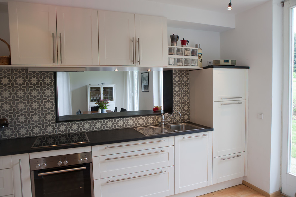 Small country single-wall eat-in kitchen in Dortmund with a drop-in sink, raised-panel cabinets, white cabinets, granite benchtops, multi-coloured splashback, cement tile splashback, stainless steel appliances, light hardwood floors and no island.
