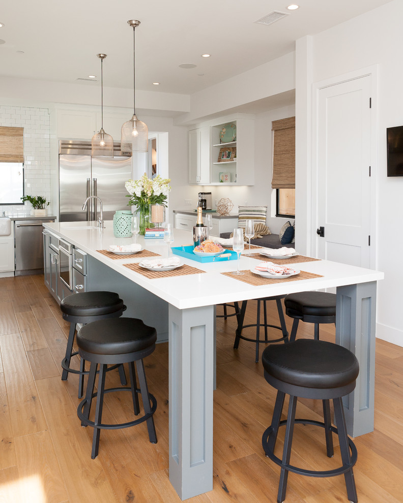 This is an example of a beach style l-shaped eat-in kitchen in Orange County with a farmhouse sink, shaker cabinets, quartzite benchtops, white splashback, subway tile splashback, stainless steel appliances, medium hardwood floors, with island and white cabinets.