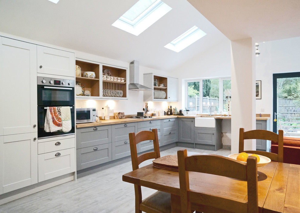 Photo of a large modern l-shaped eat-in kitchen in Buckinghamshire with a farmhouse sink, shaker cabinets, grey cabinets, wood benchtops, white splashback, stainless steel appliances, laminate floors, no island and grey floor.
