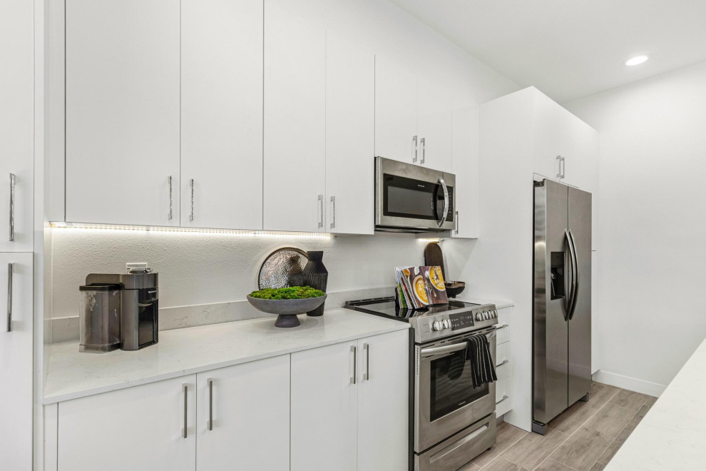 This is an example of a large contemporary u-shaped kitchen/diner in Tampa with a built-in sink, shaker cabinets, white cabinets, quartz worktops, white splashback, ceramic splashback, white appliances, light hardwood flooring, an island, brown floors and white worktops.