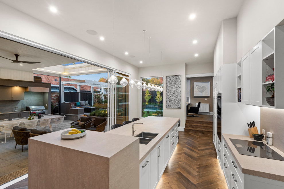 Photo of a large traditional galley open plan kitchen in Melbourne with an undermount sink, recessed-panel cabinets, white cabinets, granite benchtops, grey splashback, granite splashback, panelled appliances, medium hardwood floors, with island, brown floor and beige benchtop.