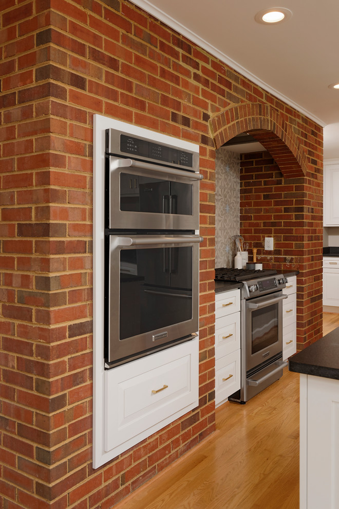 Enclosed kitchen - mid-sized transitional galley light wood floor enclosed kitchen idea in DC Metro with a farmhouse sink, raised-panel cabinets, white cabinets, solid surface countertops, gray backsplash, ceramic backsplash, stainless steel appliances and an island