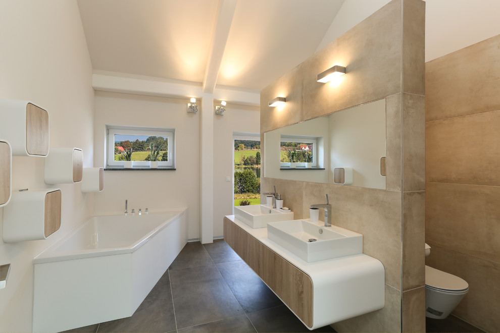 This is an example of a large contemporary bathroom in Berlin with flat-panel cabinets, light wood cabinets, a corner tub, a curbless shower, beige tile, ceramic tile, white walls, porcelain floors, a trough sink, a wall-mount toilet and solid surface benchtops.
