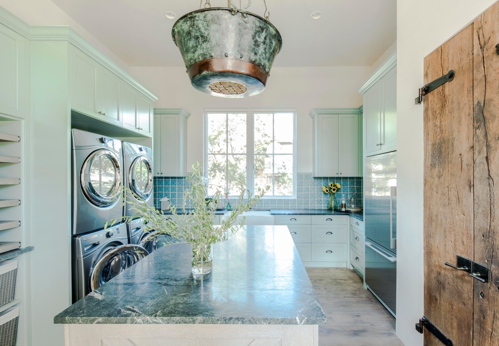 This is an example of a large country u-shaped utility room in Austin with a farmhouse sink, shaker cabinets, blue cabinets, soapstone benchtops, white walls, light hardwood floors, a stacked washer and dryer and multi-coloured benchtop.