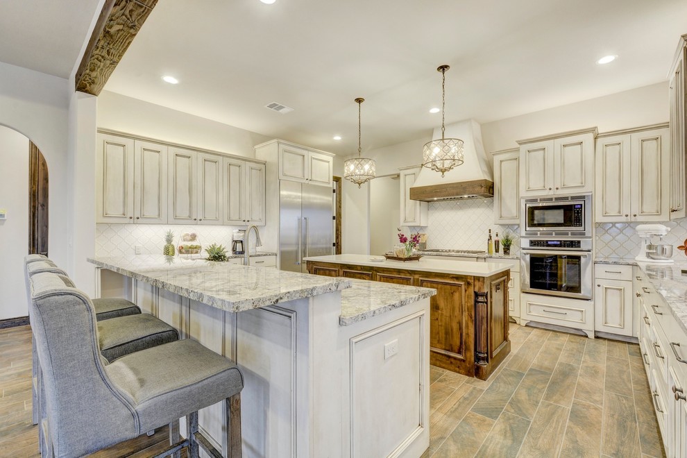 Large transitional u-shaped eat-in kitchen in Austin with an undermount sink, recessed-panel cabinets, distressed cabinets, granite benchtops, white splashback, ceramic splashback, stainless steel appliances, porcelain floors, multiple islands and brown floor.