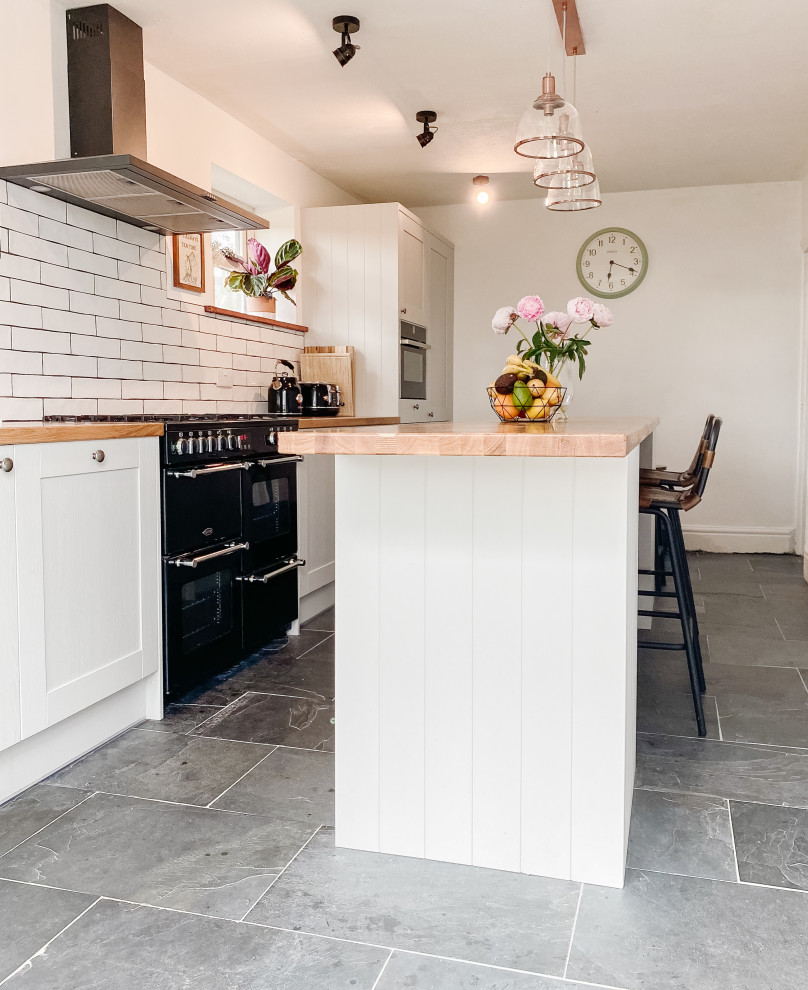 This is an example of a mid-sized traditional single-wall eat-in kitchen in Cornwall with a farmhouse sink, shaker cabinets, grey cabinets, wood benchtops, white splashback, ceramic splashback, black appliances, slate floors, with island, black floor and brown benchtop.