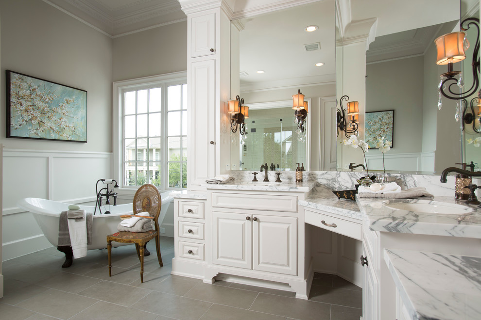 This is an example of an expansive transitional master bathroom in Houston with an undermount sink, raised-panel cabinets, white cabinets, marble benchtops, a claw-foot tub, beige walls, ceramic floors, white tile, grey benchtops, grey floor, a double vanity and a built-in vanity.