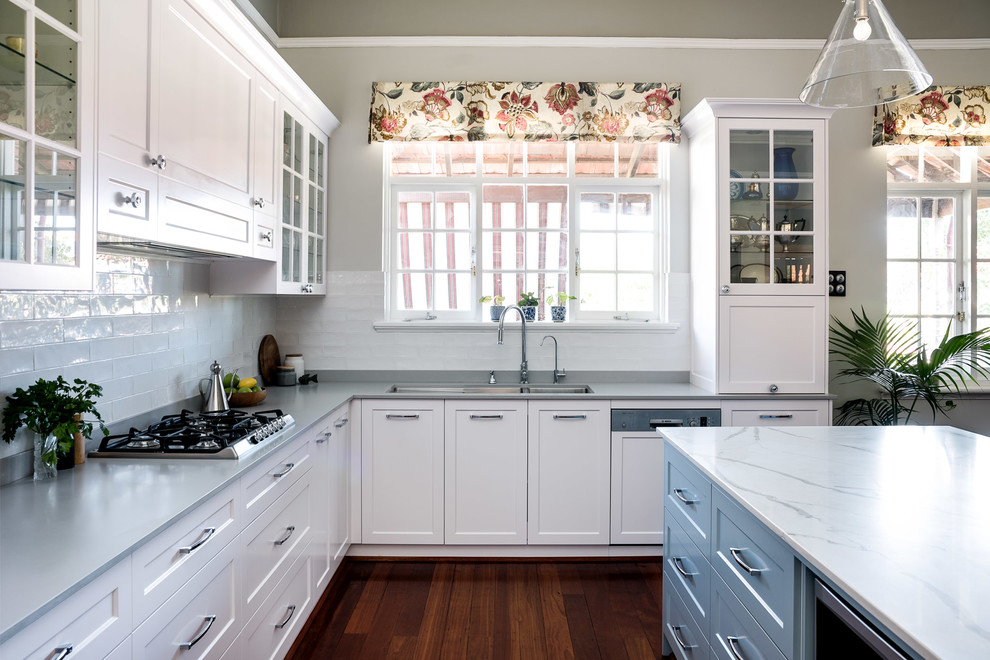 Mid-sized transitional u-shaped open plan kitchen in Perth with an undermount sink, shaker cabinets, white cabinets, quartz benchtops, white splashback, ceramic splashback, stainless steel appliances, medium hardwood floors, with island, brown floor and multi-coloured benchtop.