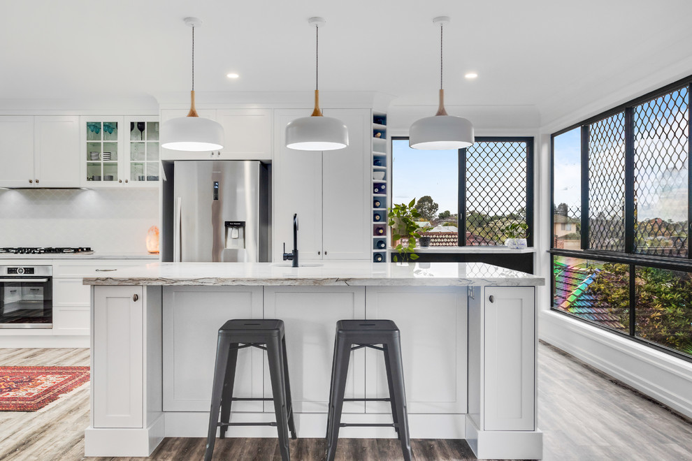 This is an example of a large transitional u-shaped eat-in kitchen in Sunshine Coast with an undermount sink, shaker cabinets, white cabinets, quartzite benchtops, white splashback, ceramic splashback, stainless steel appliances, vinyl floors, with island, white benchtop and brown floor.