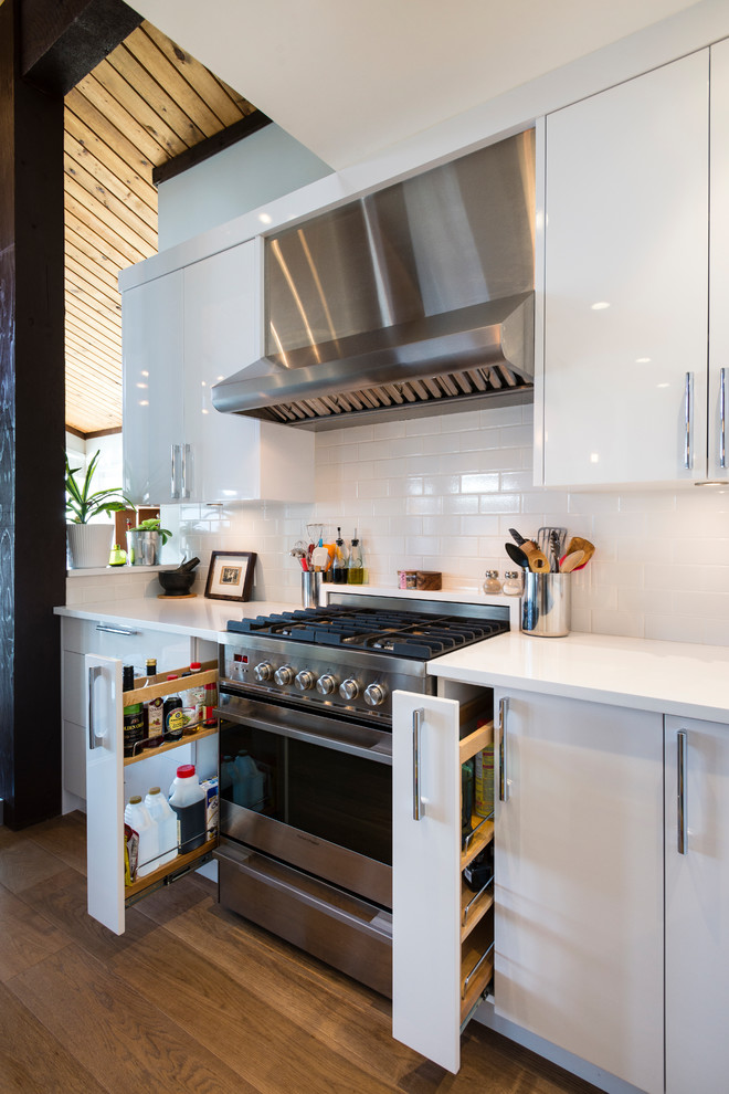 Example of a mid-sized l-shaped light wood floor and brown floor open concept kitchen design in Vancouver with an undermount sink, flat-panel cabinets, white cabinets, quartz countertops, white backsplash, ceramic backsplash, stainless steel appliances and an island