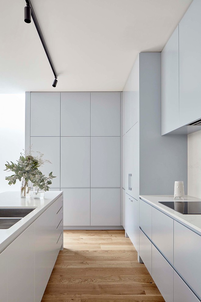 Photo of a mid-sized contemporary galley open plan kitchen in Melbourne with a double-bowl sink, flat-panel cabinets, blue cabinets, quartz benchtops, white splashback, stone slab splashback, stainless steel appliances, light hardwood floors, with island and white benchtop.