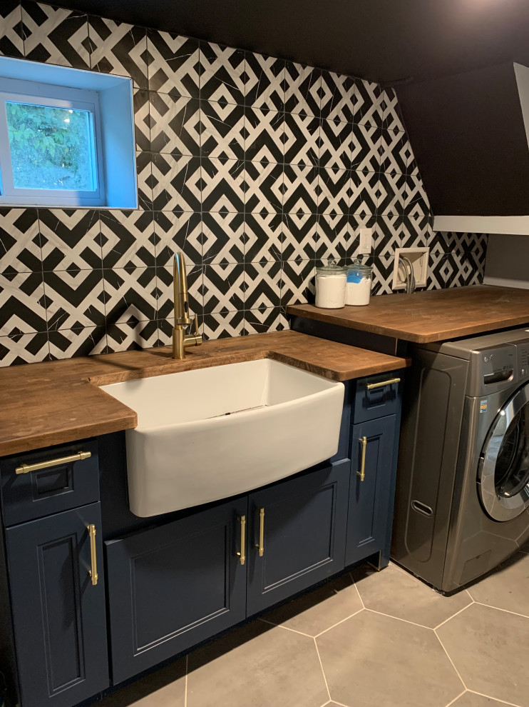 Design ideas for a medium sized contemporary galley separated utility room in New York with a belfast sink, beaded cabinets, blue cabinets, wood worktops, multi-coloured splashback, cement tile splashback, grey walls, ceramic flooring, a side by side washer and dryer, grey floors and brown worktops.