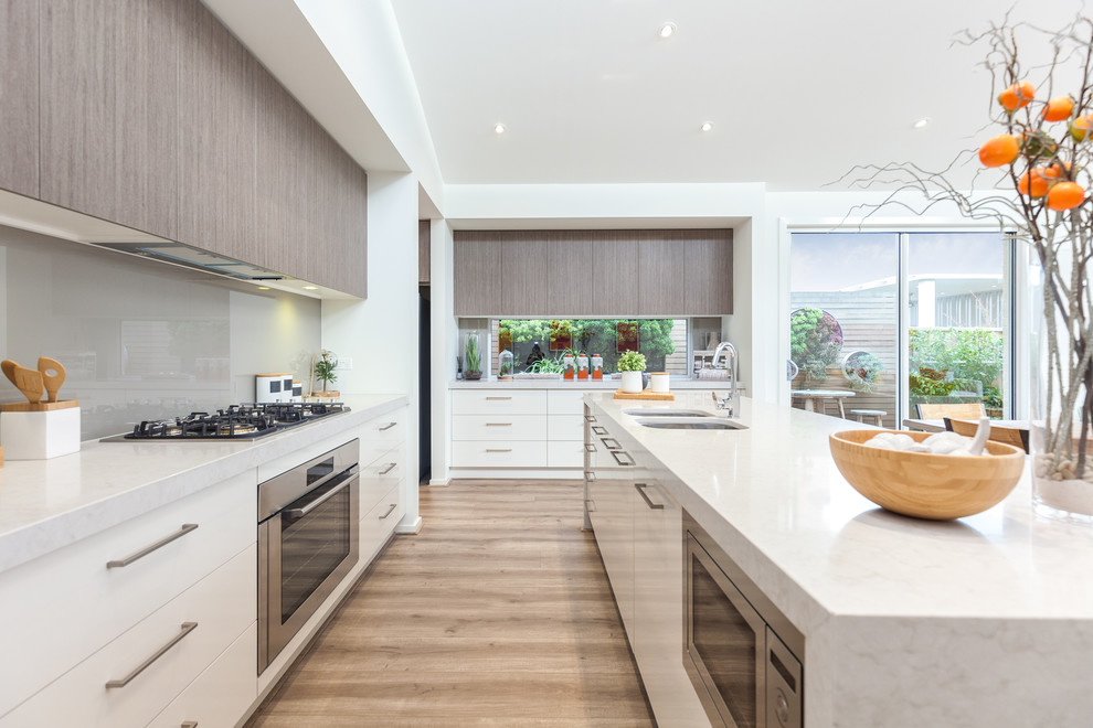 This is an example of a contemporary galley open plan kitchen in Adelaide with a double-bowl sink, flat-panel cabinets, white cabinets, marble benchtops, grey splashback, glass sheet splashback, stainless steel appliances, light hardwood floors and with island.