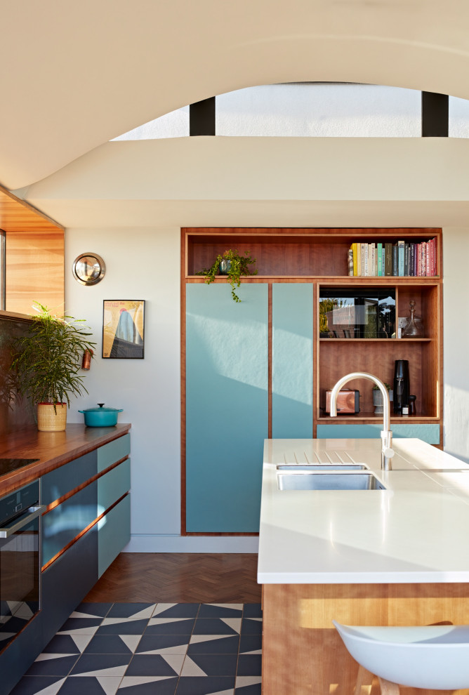 Inspiration for a large midcentury cream and black galley kitchen/diner in Sussex with a submerged sink, flat-panel cabinets, blue cabinets, composite countertops, metallic splashback, metal splashback, black appliances, ceramic flooring, an island, black floors, white worktops and a vaulted ceiling.