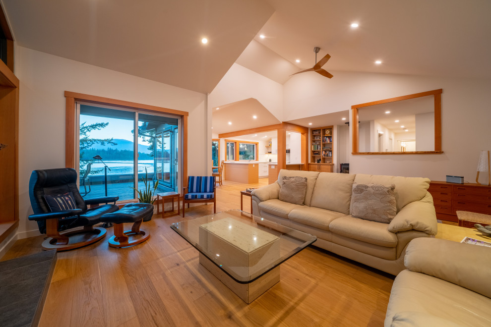 Beach style open concept family room in Vancouver with white walls, light hardwood floors, a standard fireplace, a brick fireplace surround, a wall-mounted tv, brown floor, vaulted and panelled walls.