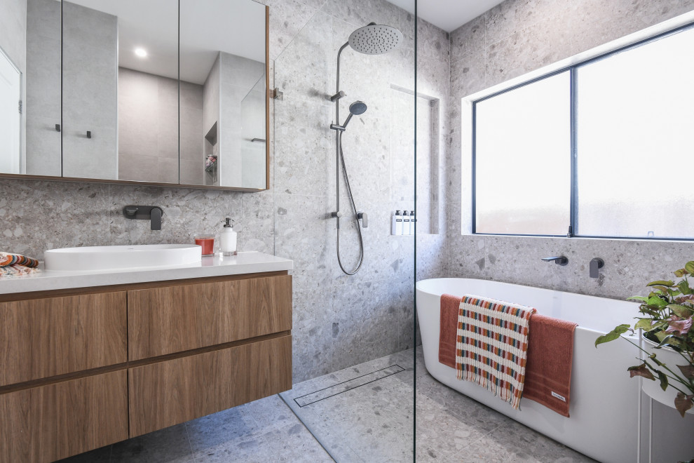 Foto på ett funkis vit badrum, med ett fristående badkar, grå kakel, ett fristående handfat, bänkskiva i kvarts, grått golv, med dusch som är öppen, släta luckor, skåp i mellenmörkt trä, en kantlös dusch och grå väggar