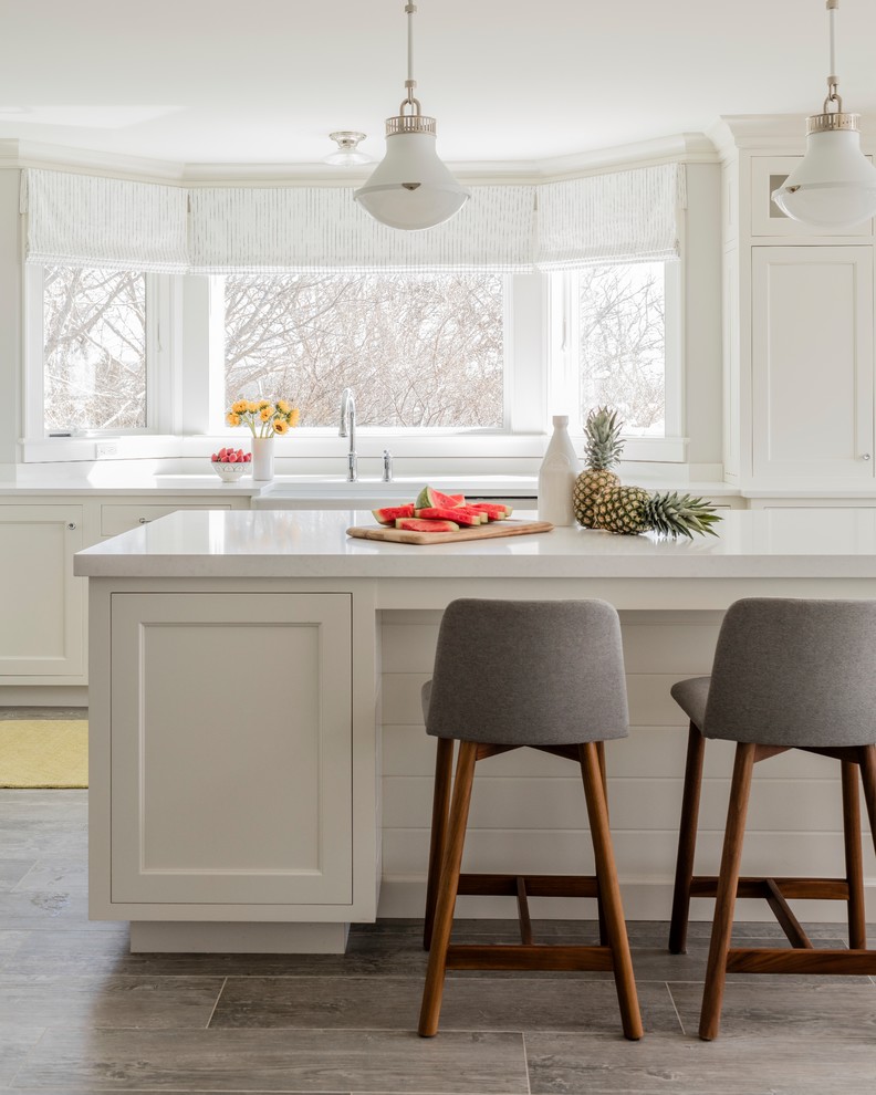 This is an example of a large beach style eat-in kitchen in New York with a farmhouse sink, shaker cabinets, white cabinets, quartz benchtops, white splashback, ceramic splashback, stainless steel appliances, porcelain floors, with island, brown floor and white benchtop.