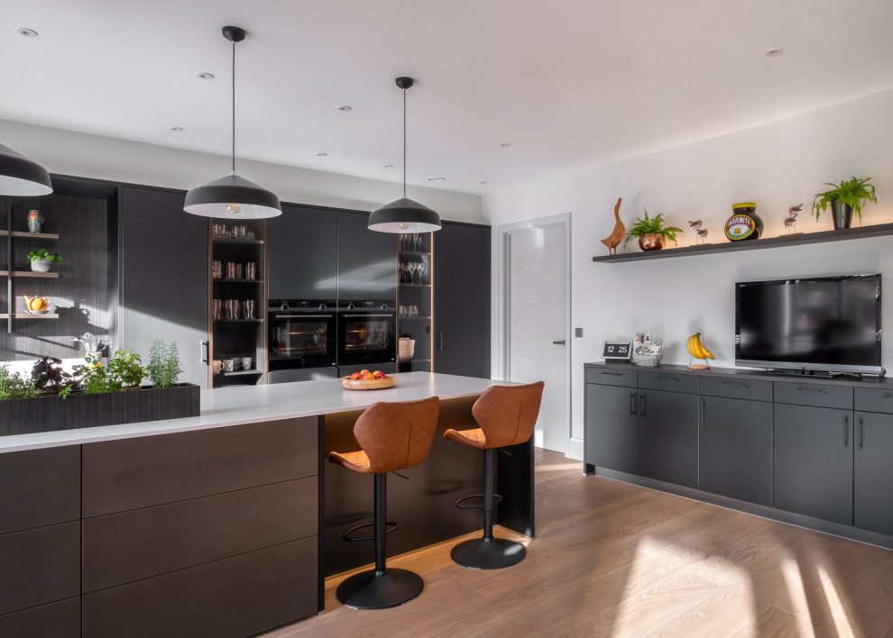 This is an example of a large contemporary eat-in kitchen in Gloucestershire with a drop-in sink, flat-panel cabinets, black cabinets, solid surface benchtops, white splashback, black appliances, with island and white benchtop.