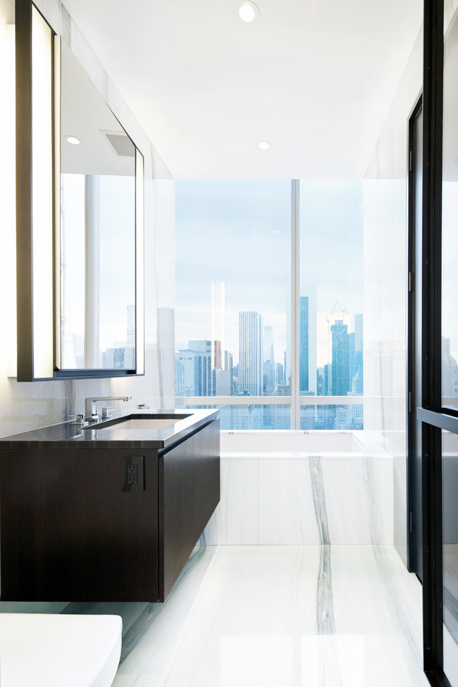 Modern bathroom in New York with furniture-like cabinets, dark wood cabinets, marble benchtops, an undermount tub, white tile, stone slab, white walls and marble floors.