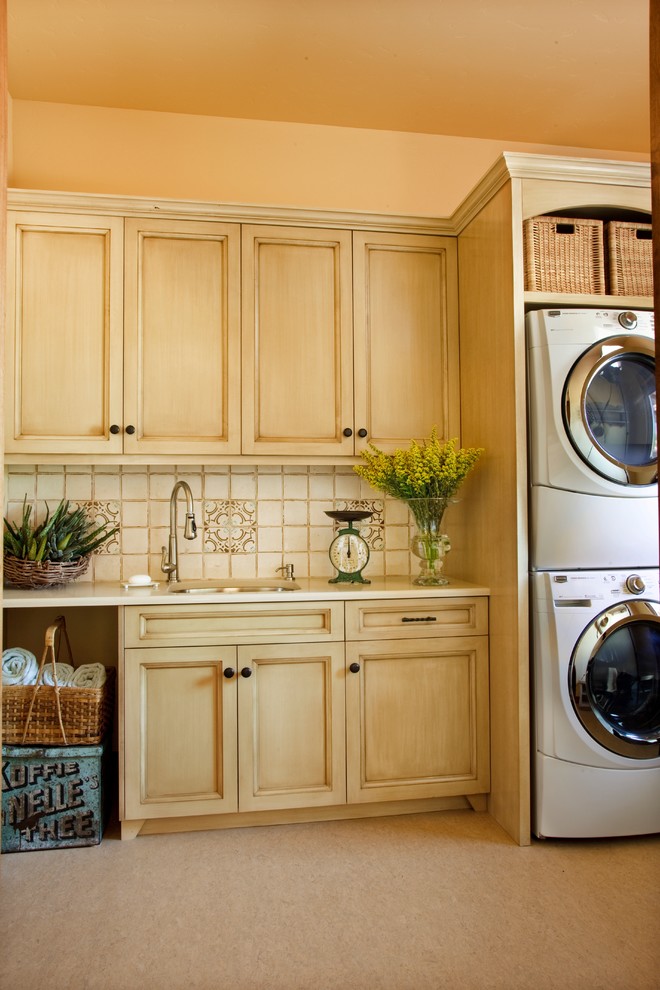 Photo of a mid-sized mediterranean single-wall dedicated laundry room in Other with an undermount sink, recessed-panel cabinets, light wood cabinets, quartz benchtops, carpet, a stacked washer and dryer, beige floor, beige benchtop and beige walls.