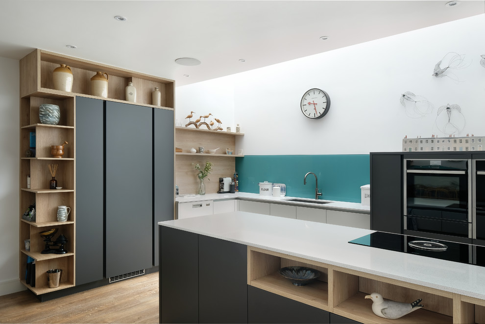 This is an example of a mid-sized contemporary l-shaped eat-in kitchen in Gloucestershire with flat-panel cabinets, grey cabinets, quartzite benchtops, blue splashback, glass sheet splashback, black appliances, medium hardwood floors, white benchtop, an undermount sink and a peninsula.